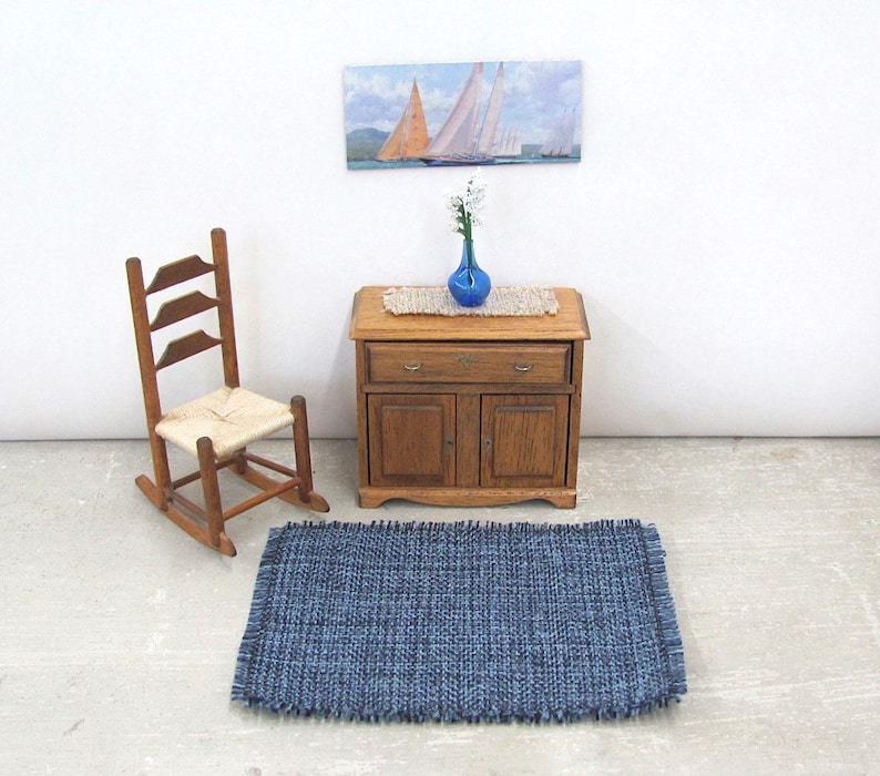 Inch to Foot Dollhouse Miniature Rug, 1:12 Scale Woven Small Indigo Navy Blue and Light Blue Floor Carpet for Modern Coastal Seaside Cottage or Traditional Rustic Nautical Beach Doll House. Styled with a rocking chair, cabinet with a linen runner and blue glass vase with white flowers, a picture of racing sailing ships on the wall.