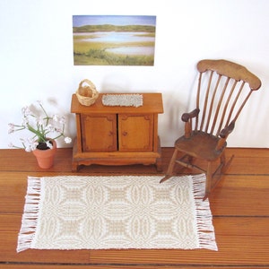 May include: A miniature dollhouse room with a wooden rocking chair, a small wooden cabinet with two doors, a woven rug, a basket, and a framed landscape painting on the wall.
