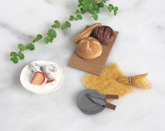 May include: A miniature scene with a white plate containing a red and white cheese wedge, two small white cheese balls, and a small brown cheese wedge. There is a brown cardboard tray with three miniature loaves of bread, one brown, one white, and one long loaf. There is a small grey metal pan with a wooden handle, a yellow cloth, and a small brown hessian bag.