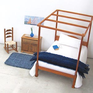 Inch to Foot Dollhouse Miniature Rug, 1:12 Scale Woven Small Indigo Navy Blue and Light Blue Floor Carpet for Modern Coastal Seaside Cottage or Traditional Rustic Nautical Beach Doll House. Styled with a rocking chair to the left; in front of a cabinet with a linen runner and blue glass vase with white flowers; a four post bed with white bedspread and blue blanket, a book and glasses to the right; a picture of racing sailing ships on the wall.