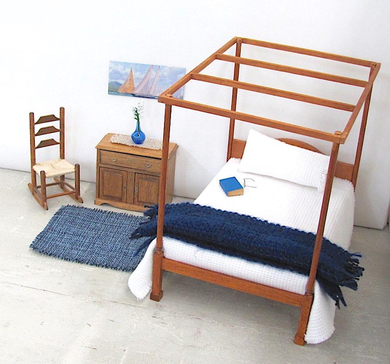 Inch to Foot Dollhouse Miniature Rug, 1:12 Scale Woven Small Indigo Navy Blue and Light Blue Floor Carpet for Modern Coastal Seaside Cottage or Traditional Rustic Nautical Beach Doll House. Styled with a rocking chair to the left; in front of a cabinet with a linen runner and blue glass vase with white flowers; a four post bed with white bedspread and blue blanket, a book and glasses to the right; a picture of racing sailing ships on the wall.