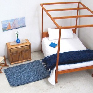 Inch to Foot Dollhouse Miniature Rug, 1:12 Scale Woven Small Indigo Navy Blue and Light Blue Floor Carpet for Modern Coastal Seaside Cottage or Traditional Rustic Nautical Beach Doll House. Styled with a rocking chair to the left; in front of a cabinet with a linen runner and blue glass vase with white flowers; a four post bed with white bedspread and blue blanket, a book and glasses to the right; a picture of racing sailing ships on the wall.