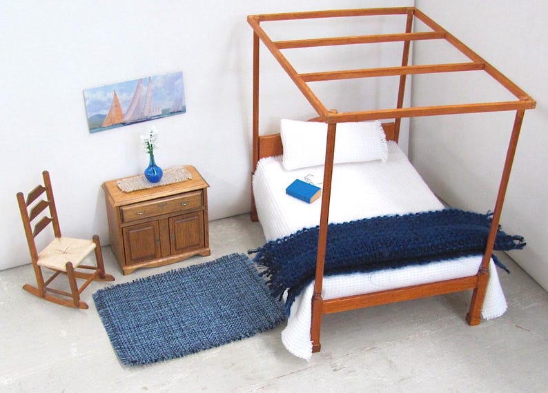 Inch to Foot Dollhouse Miniature Rug, 1:12 Scale Woven Small Indigo Navy Blue and Light Blue Floor Carpet for Modern Coastal Seaside Cottage or Traditional Rustic Nautical Beach Doll House. Styled with a rocking chair to the left; in front of a cabinet with a linen runner and blue glass vase with white flowers; a four post bed with white bedspread and blue blanket, a book and glasses to the right; a picture of racing sailing ships on the wall.