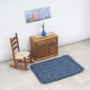 Inch to Foot Dollhouse Miniature Rug, 1:12 Scale Woven Small Indigo Navy Blue and Light Blue Floor Carpet for Modern Coastal Seaside Cottage or Traditional Rustic Nautical Beach Doll House. Styled with a rocking chair, cabinet with a linen runner and blue glass vase with white flowers, a picture of racing sailing ships on the wall.
