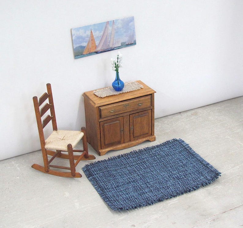 Inch to Foot Dollhouse Miniature Rug, 1:12 Scale Woven Small Indigo Navy Blue and Light Blue Floor Carpet for Modern Coastal Seaside Cottage or Traditional Rustic Nautical Beach Doll House. Styled with a rocking chair, cabinet with a linen runner and blue glass vase with white flowers, a picture of racing sailing ships on the wall.