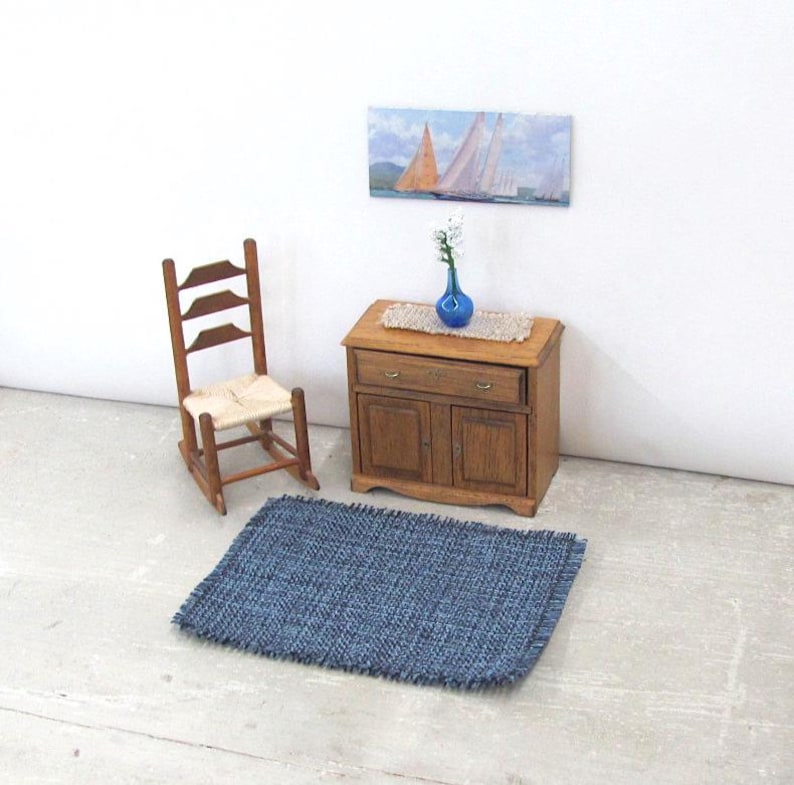 Inch to Foot Dollhouse Miniature Rug, 1:12 Scale Woven Small Indigo Navy Blue and Light Blue Floor Carpet for Modern Coastal Seaside Cottage or Traditional Rustic Nautical Beach Doll House. Styled with a rocking chair, cabinet with a linen runner and blue glass vase with white flowers, a picture of racing sailing ships on the wall.