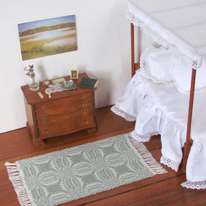 Handmade, Handcrafted 1:12 Scale Dollhouse Miniature Rug, Artisan Hand Woven Jade Green Carpet for Traditional Country Farmhouse or Modern Coastal Beach Cottage Decor is a perfect Doll House Furniture Accessory for your Miniature Bedroom or Living Room! Shown in a Bedroom scene with white canopy bed with a white ruffled bed skirt and bedspread, wooden dresser with  a small mirror, a book, and a few decorative items. A framed picture of a landscape is on the wall behind the dresser.