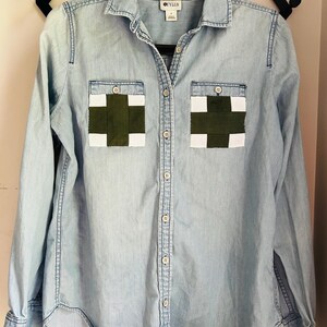 May include: Light blue denim shirt with two chest pockets featuring a green and white cross design. The shirt has long sleeves, a button-down front, and the brand name "CYLUS" is visible on the tag.
