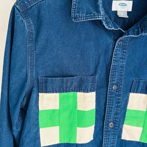 May include: A blue denim shirt with two front pockets featuring a green and white patchwork design. The shirt has a button-down front, long sleeves, and a collar. The Old Navy brand label is visible.
