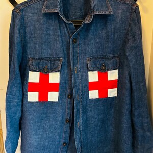 May include: A denim shirt with two front pockets, each featuring a white rectangle with a red cross. The shirt has a button-down front and long sleeves. The shirt is a medium blue color.