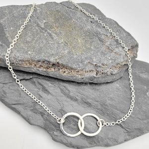 May include: A silver-colored necklace featuring a delicate chain and two interlocking circular pendants. The metal rings have a slightly hammered, textured finish, creating a reflective surface that catches the light along the thin, circular links.
