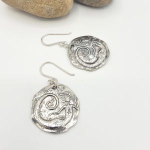 May include: A pair of silver-toned earrings. Each earring features a circular, textured pendant with a mermaid design. The mermaid is depicted with flowing hair and a swirling tail. The earrings have a simple hook closure.