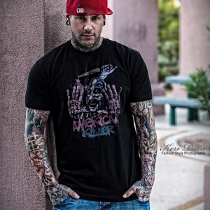 May include: A man with tattoos on his arms is wearing a black t-shirt with a graphic print of a skull and the words "American Killer". He is also wearing a red baseball cap, blue jeans, and a silver earring.