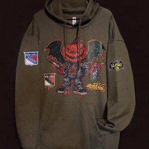 May include: Olive green hoodie with a large graphic of a pumpkin-headed character holding a knife and spray paint. Patches include "New York Rangers" and "Halfway Toons". The design features red, black, and green accents.