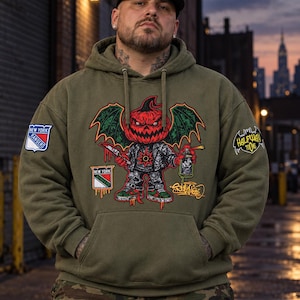 May include: An olive green hooded sweatshirt featuring a graphic of a winged pumpkin character holding a knife and a spray paint can. The hoodie includes a New York Rangers logo on the right sleeve and a Halfway Toons logo on the left sleeve.
