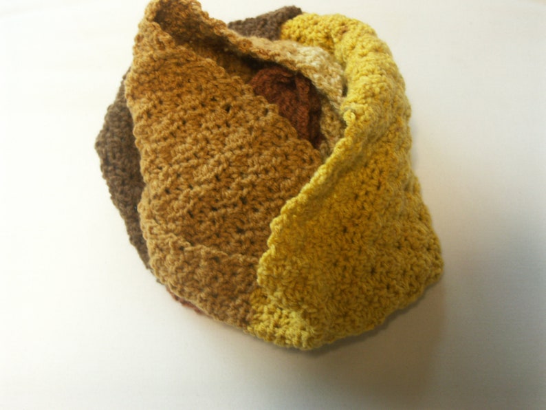 May include: A crocheted scarf in shades of brown, yellow, and gold. The scarf is made of a soft, textured yarn and is folded into a decorative shape.