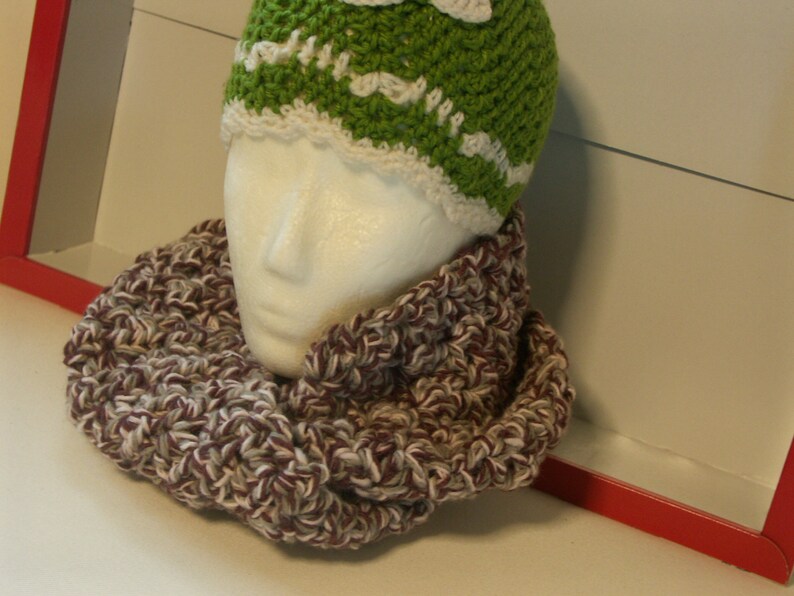 May include: A green and white crocheted beanie hat and a matching infinity scarf. The scarf is a mix of brown, white, and burgundy yarn. The hat is on a mannequin head.