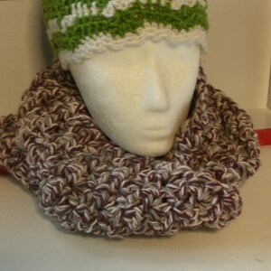 May include: A handmade crochet infinity scarf in burgundy, white, and grey, displayed on a mannequin head. A green and white crochet hat is visible above the scarf. The scarf is thick and textured.