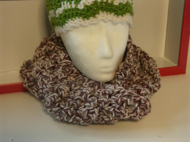 May include: A handmade crochet infinity scarf in burgundy, white, and grey, displayed on a mannequin head. A green and white crochet hat is visible above the scarf. The scarf is thick and textured.