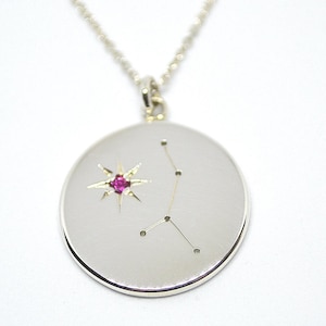 May include: A silver necklace with a round pendant featuring a constellation design and a pink gemstone in the center.