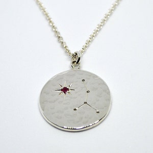 May include: A silver necklace with a round pendant featuring a constellation design and a red gemstone in the center. The pendant has a hammered texture.