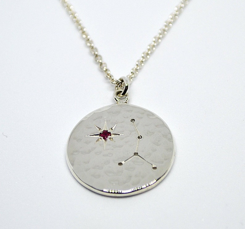 May include: A silver necklace with a round pendant featuring a constellation design and a red gemstone in the center. The pendant has a hammered texture.