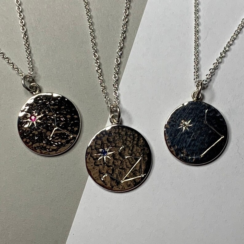 May include: Three silver necklaces with round pendants featuring a constellation design. Each pendant has a small gemstone in the center, one is pink, one is blue, and one is clear.