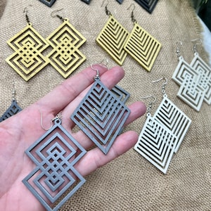 Puede incluir: Un conjunto de pendientes de madera geométricos en varios colores, incluyendo dorado, gris y blanco. Los pendientes tienen forma de cuadrados con intrincados patrones.