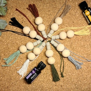 May include: Wooden bead tassels in a variety of colours, such as brown, black, green, and yellow, arranged in a circular pattern. Two essential oil bottles are also present, one labelled "Ylang-Ylang" and the other "Peace".