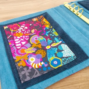 May include: A blue denim binder with a colourful floral fabric pocket on the front. The binder has a gold metal ring mechanism.