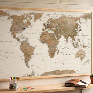 May include: A rectangular world map in a light wood frame with a cream background and brown continents. The map features various coloured push pins marking locations across the globe. It is displayed on a wall above a wooden shelf with accessories.