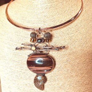 May include: A copper-colored choker necklace with a pendant featuring a large, oval, brown and black striped stone. The pendant has a unique design with smaller stones and decorative elements. The necklace is displayed on a neutral-toned background.