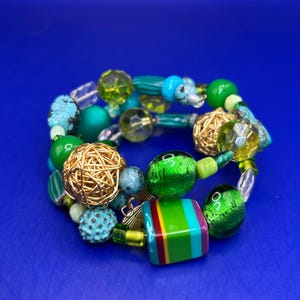 May include: A colorful beaded bracelet featuring a variety of beads in shades of green, turquoise, and gold. The bracelet includes round, faceted, and textured beads, with a rectangular bead with colorful stripes. The bracelet is on a blue background.