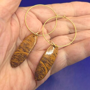 May include: A pair of gold hoop earrings with elongated, patterned stone pendants. The pendants feature a mix of brown and yellow tones, creating a marbled effect. The earrings are displayed against a neutral background.