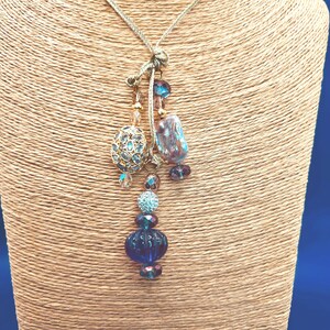 May include: A gold-toned necklace with a pendant featuring a cluster of blue and purple beads. The pendant includes a filigree oval, a large blue bead, and other decorative elements. The necklace is displayed on a tan mannequin.