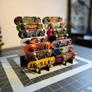 Pode incluir: Uma coleção de mini-skates, cada um com gráficos e designs coloridos únicos. Os skates são exibidos em suportes pretos, mostrando a variedade de obras de arte. Um skate amarelo tem o nome "JOSL" impresso.