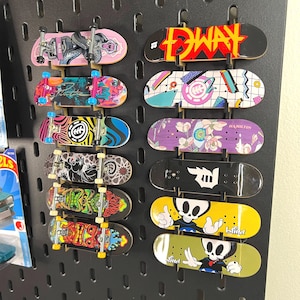 Puede incluir: Una colección de doce monopatines para dedos montados en un panel perforado negro. Las tablas presentan varios diseños gráficos, incluyendo calaveras, patrones florales, formas abstractas y logotipos. Algunas tablas tienen ejes de metal.