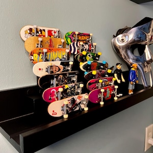 May include: A black shelf with a collection of colourful fingerboard skateboards mounted on it. The skateboards have various designs, including a Baltimore Orioles logo, a skull, and a cartoon character. There are also two action figures on the shelf.
