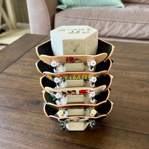 May include: A stack of five miniature skateboards with black grip tape and colourful graphics. The skateboards are supported by a grey, geometric block. The decks feature various designs, including text and illustrations.