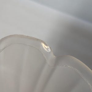 May include: Close-up of a clear, scalloped-edge glass bowl. The bowl has a frosted appearance and a slightly irregular rim. The light reflects off the surface, highlighting the curves and texture of the glass.