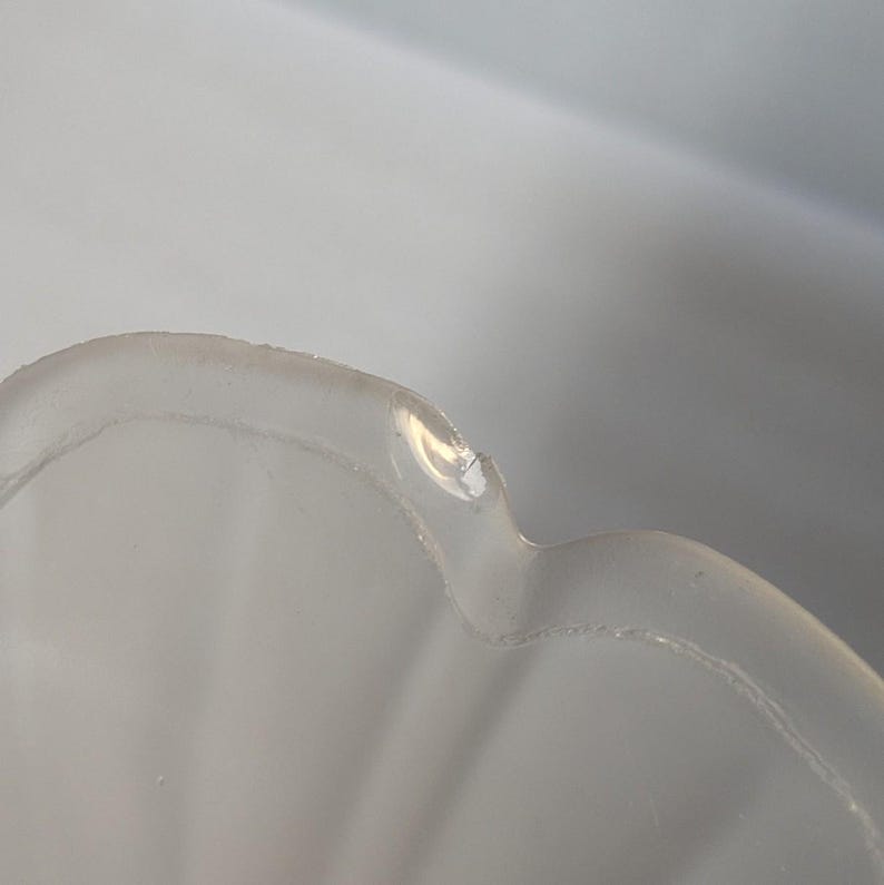 May include: Close-up of a clear, scalloped-edge glass bowl. The bowl has a frosted appearance and a slightly irregular rim. The light reflects off the surface, highlighting the curves and texture of the glass.