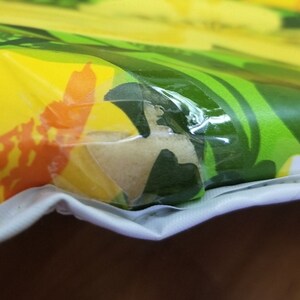 Pode incluir: Close-up de um tecido estampado em amarelo, laranja e verde coberto com uma pel&iacute;cula de pl&aacute;stico transparente. O tecido est&aacute; dobrado e o pl&aacute;stico est&aacute; ligeiramente enrugado.