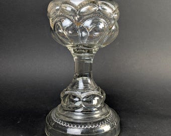 May include: Clear glass oil lamp with a decorative, rounded body and base. The lamp features a detailed, embossed pattern and a metal top. The design includes a stem and a circular base, showcasing a vintage aesthetic.