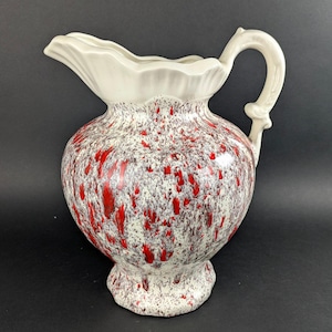 May include: A vintage ceramic pitcher with a white, ruffled rim and handle. The body features a speckled pattern of red and brown against a cream background. The pitcher has a bulbous shape and a pedestal base.