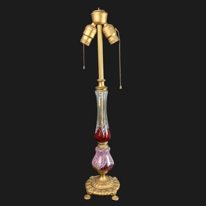 May include: An antique floor lamp with a gold-colored metal base and accents. The lamp features a clear glass column with red and pink accents, and three light sockets at the top. The lamp is a decorative lighting fixture.