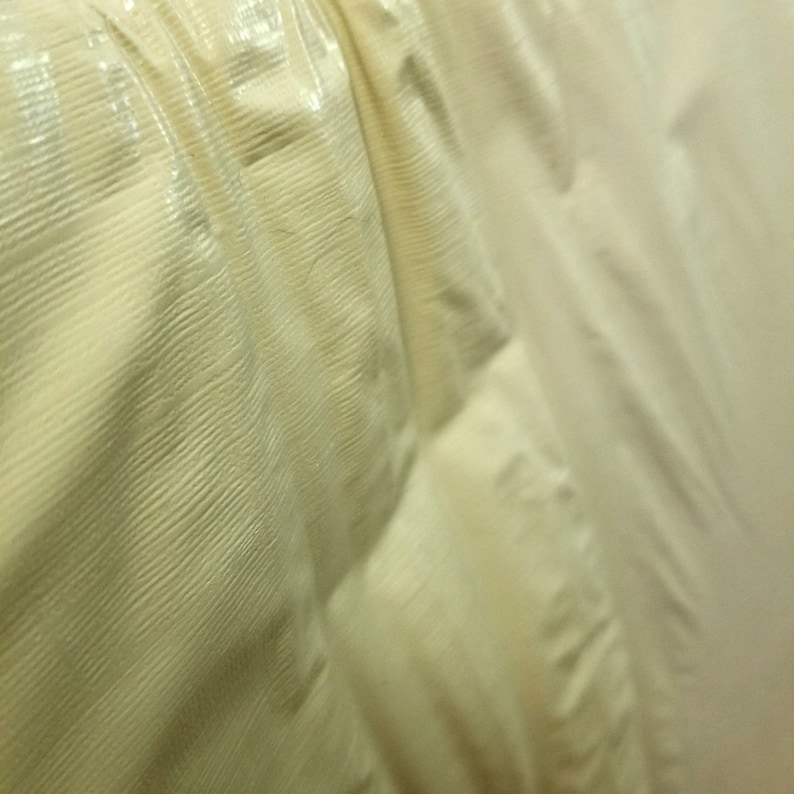 Pode incluir: Close-up de um tecido cor de creme com um padr&atilde;o acolchoado texturizado. O tecido tem um brilho sutil e parece ser feito de um material leve.