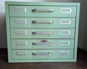 May include: A vintage green metal cabinet with five drawers. Each drawer has a silver handle and a label holder. The cabinet is set on a dark wooden surface.