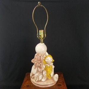 May include: A ceramic table lamp featuring a whimsical design of a rag doll holding a ball. The lamp is painted in shades of white, brown, and yellow. The lampshade is missing.