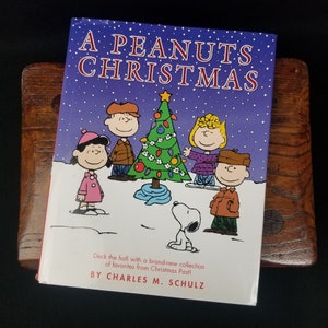 May include: A Peanuts Christmas book cover featuring a purple background with white snowflakes, a decorated Christmas tree, and the Peanuts characters Charlie Brown, Lucy, Linus, and Snoopy. The book title is "A Peanuts Christmas" in white text.