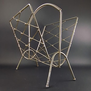 May include: A gold-toned metal magazine rack with a geometric design and a curved handle. The rack has three legs and is made of wire.